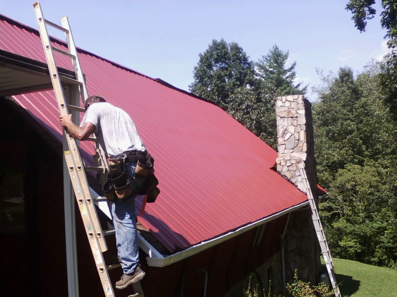 Roofer on a ladder beside a red metal roof and stone chimney — Soffit & Fascia Repair by Roofing Brothers in Merrill